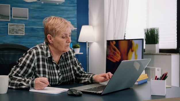 Senior stylish woman taking notes in notebook while using laptop at home. Old freelancer writing details on book while working from workspace in living room, checking financial project of company