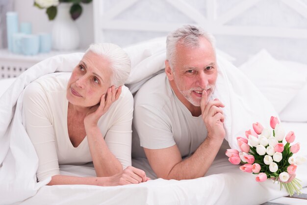 Senior thoughtful woman lying on bed beside her husband doing silence gesture holding tulip flower bouquet