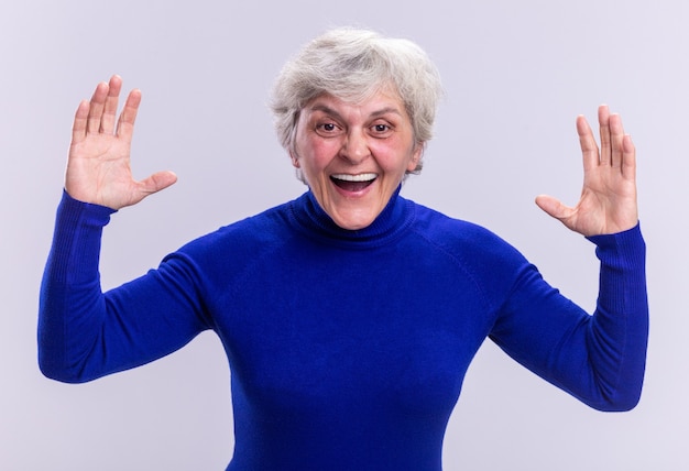 Foto gratuita donna anziana in dolcevita blu che guarda la telecamera felice ed eccitata con le braccia alzate in piedi sopra il bianco