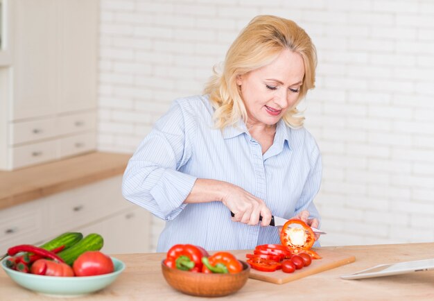 Senior woman cutting the slices of red bell pepper with knife on chopping board in the kitchen