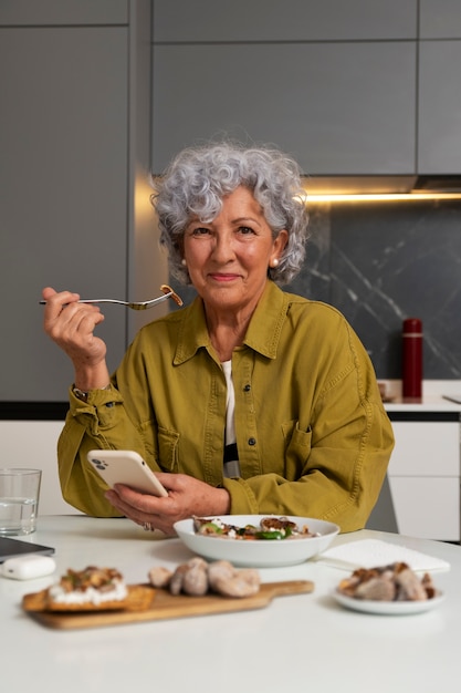 Foto gratuita donna anziana che mangia il piatto di fichi a casa e utilizza lo smartphone