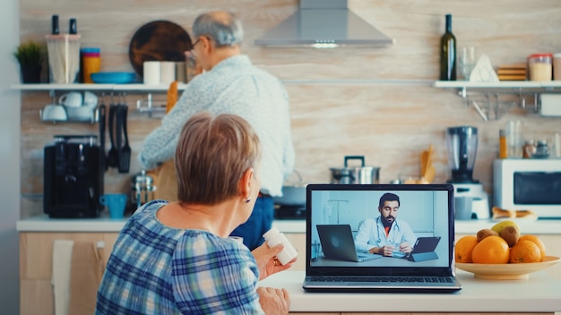 Senior woman holding bottle of pills during video conference with doctor using laptop in kitchen. Online health consultation for elderly people drugs ilness advice on symptoms, physician telemedicine