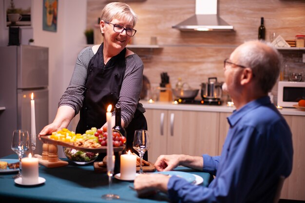 Старшая женщина держит деревянную тарелку с мужем и смотрит на него во время праздничного ужина. Пожилая пара разговаривает, сидя за столом на кухне, наслаждаясь едой, празднуя годовщину.