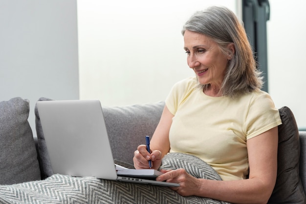 Старшая женщина дома на диване, используя ноутбук и делая заметки