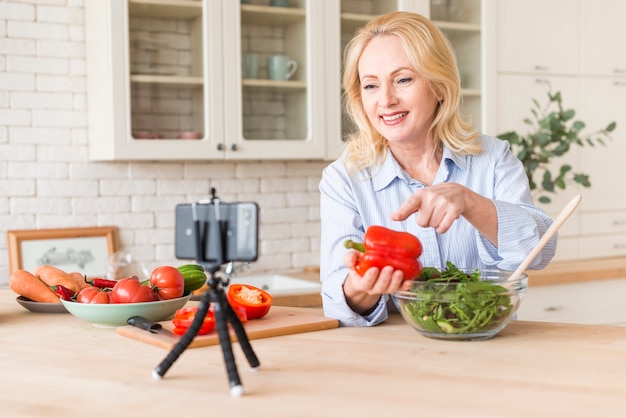 Foto gratuita donna senior che fa videochiamata sul telefono cellulare che mostra peperone mentre preparando insalata