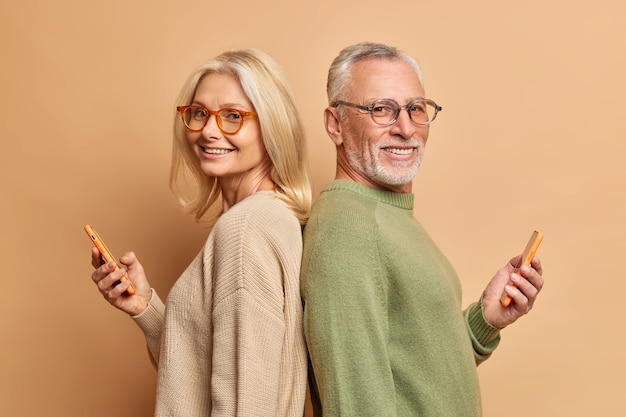 Senior woman and man stand backs to each other use modern cellphones stand backs to each other wear spectacles and casual sweaters isolated over brown wall