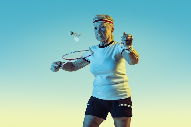Senior woman playing badminton in sportwear on gradient background in neon light