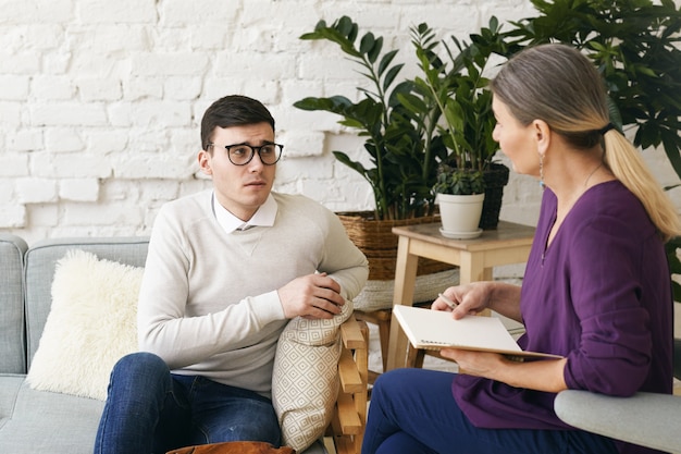 Senior woman psychotherapist or counselor writing something down in notebook during therapy session with frustrated depressed young male in spectacles. Psychology, counseling and mental health
