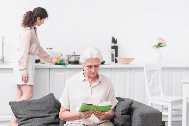 Senior woman reading book in front of woman doing housekeeping work