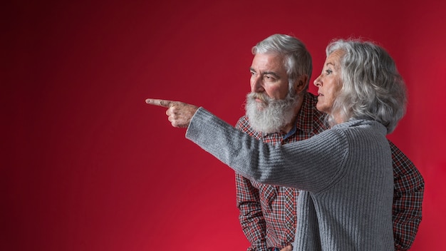 Free photo senior woman showing something to her husband by pointing finger against red background