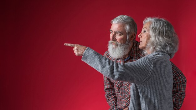 Senior woman showing something to her husband by pointing finger against red background