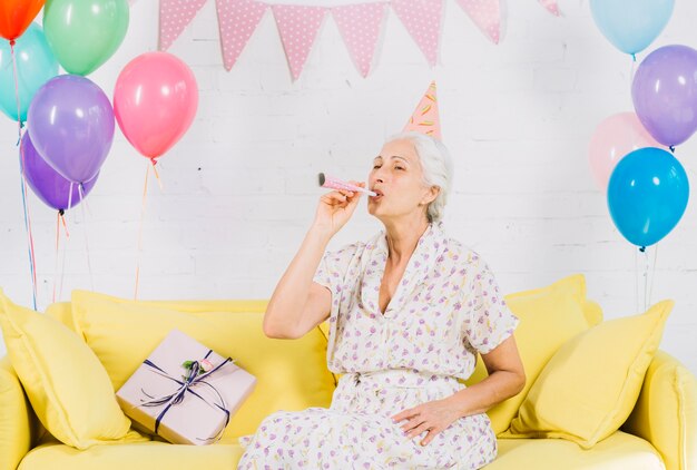 Senior woman sitting on sofa blowing party horn