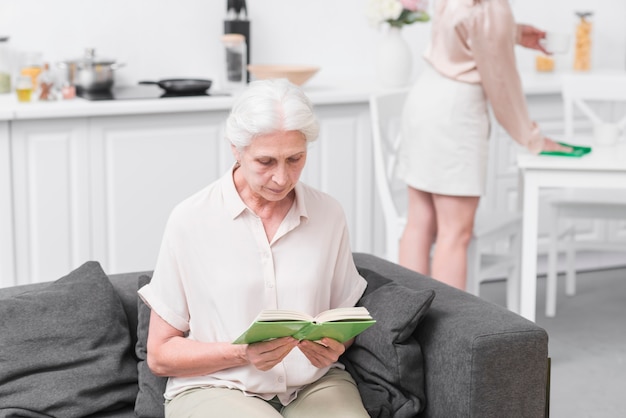 Старший женщина, сидя на диване, чтение книги у себя дома