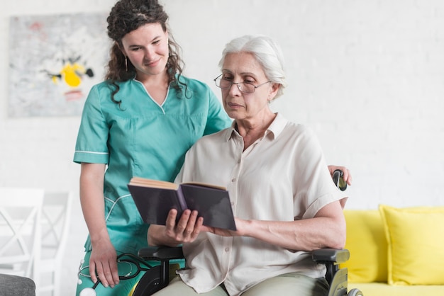 Senior woman sitting on wheelchair reading book with nurse standing behind