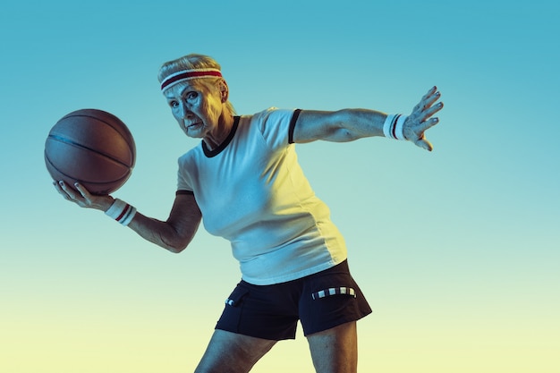 Foto gratuita senior donna in abbigliamento sportivo giocando a basket su sfondo sfumato, luce al neon. il modello femminile in ottima forma rimane attivo. concetto di sport, attività, movimento, benessere, fiducia. copyspace.