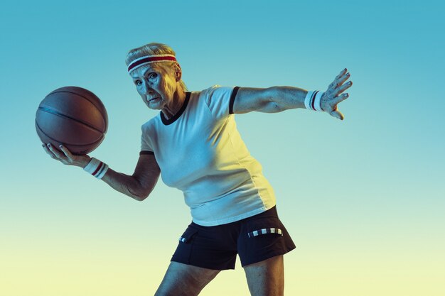 Старшая женщина в спортивной одежде играет в баскетбол на градиентном фоне, неоновый свет. Девушка-модель в отличной форме остается активной. Понятие спорта, активности, движения, благополучия, уверенности. Copyspace.