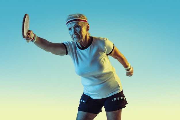 Senior woman in sportwear training in table tennis on gradient background, neon light. Female model in great shape stays active. Concept of sport, activity, movement, wellbeing, confidence. Copyspace.