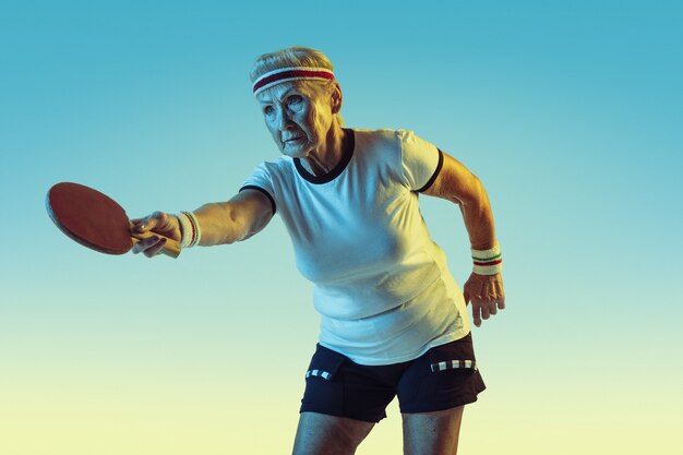 Senior woman in sportwear training in table tennis on gradient background, neon light.