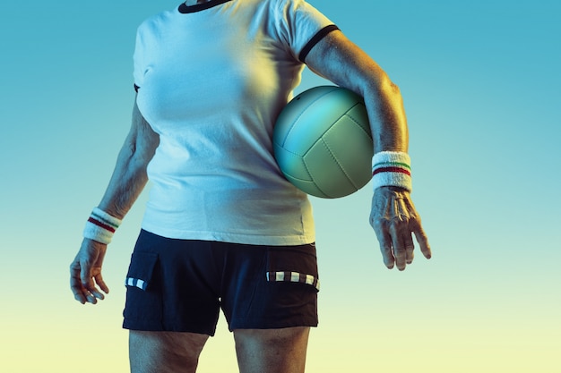Senior woman in sportwear training in volleyball on gradient background, neon light. Female model in great shape stays active. Concept of sport, activity, movement, wellbeing, confidence. Copyspace.
