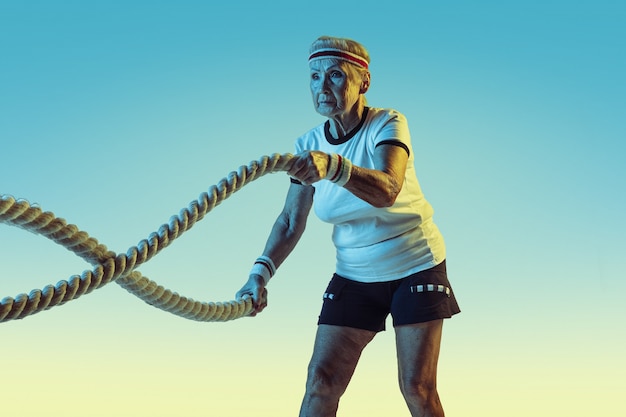 Free photo senior woman in sportwear training with ropes on gradient background, neon light. female model in great shape stays active. concept of sport, activity, movement, wellbeing, confidence. copyspace.
