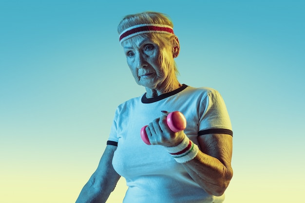 Free photo senior woman in sportwear training with weights on gradient background, neon light. female model in great shape stays active. concept of sport, activity, movement, wellbeing, confidence. copyspace.