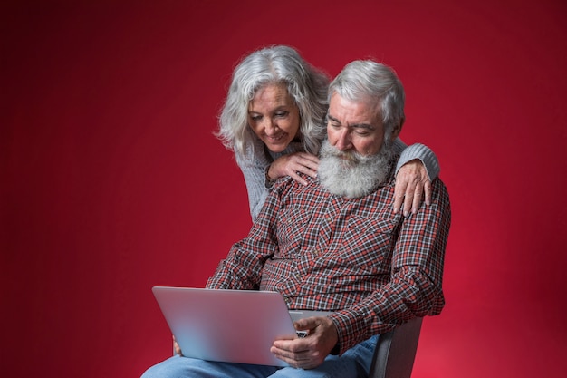 Donna senior che sta dietro il suo uomo che si siede sulla sedia che esamina computer portatile contro il fondo rosso