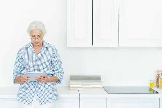 Senior woman standing in kitchen using digital tablet