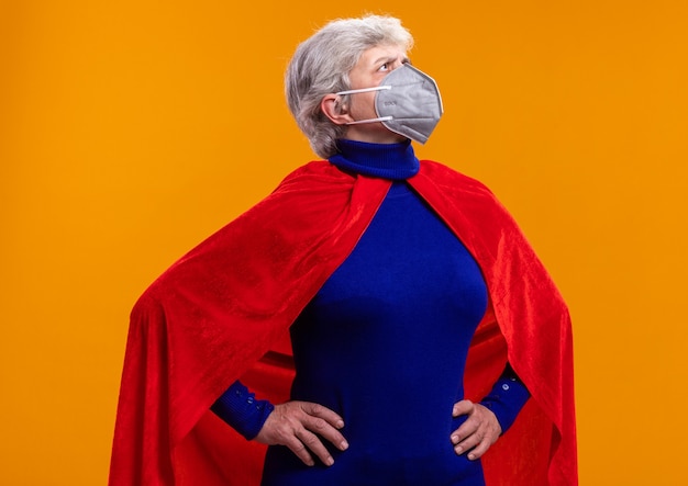 Senior woman superhero wearing red cape and facial protective mask looking aside