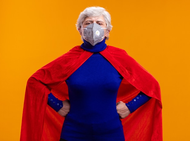 Senior woman superhero wearing red cape and facial protective mask looking at camera with confident expression with arms at hip standing over orange background