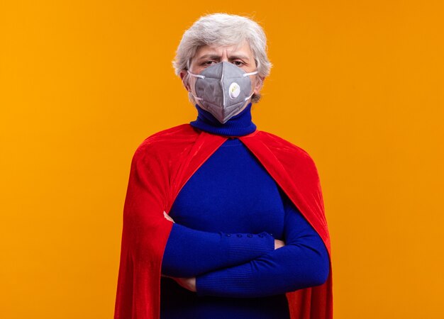 Senior woman superhero wearing red cape and facial protective mask looking at camera with serious confident expression standing over orange background