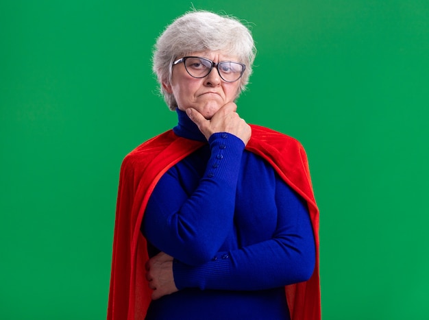 Free photo senior woman superhero wearing red cape and glasses looking at camera with skeptic expression standing over green background