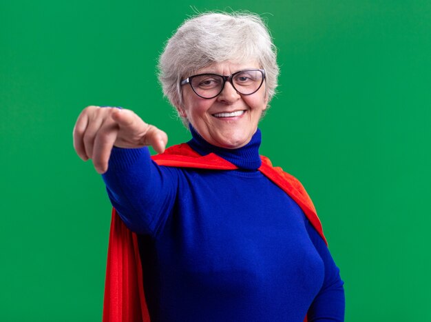 Senior woman superhero wearing red cape and glasses pointing with index finger at camera