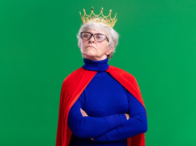 Senior woman superhero wearing red cape and glasses with crown on head looking aside