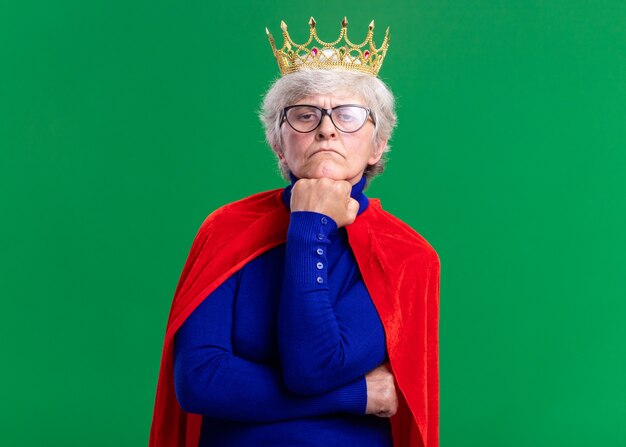 Старшая женщина-супергерой в красной накидке и очках с короной на голове, смотрящая в камеру