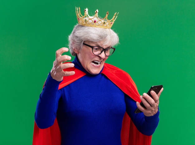 Senior woman superhero wearing red cape and glasses with crown on head looking at screen of her smartphone screaming with aggressive expression standing over green background