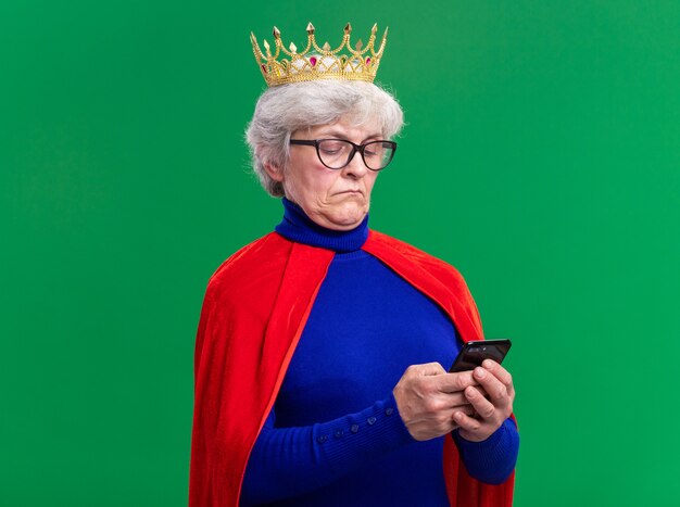 緑の背景の上に立って自信を持って見えるスマートフォンを使用して頭に王冠と赤いマントと眼鏡を身に着けている年配の女性のスーパーヒーロー