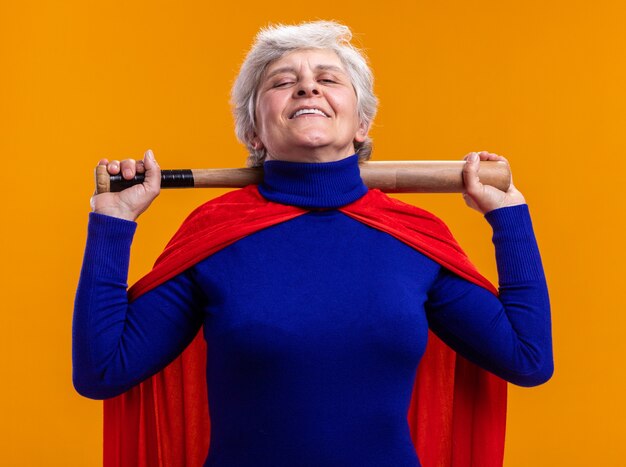 Senior woman superhero wearing red cape holding baseball bat looking at camera pleased and confident standing over orange background