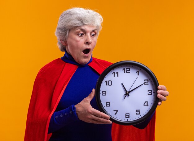 Senior woman superhero wearing red cape holding clock looking at it amazed and surprised 