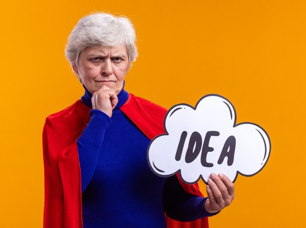 Senior woman superhero wearing red cape holding speech bubble sign with word idea looking at camera with pensive expression on face thinking standing over orange background