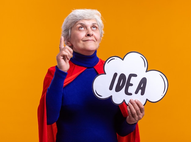 Senior woman superhero wearing red cape holding speech bubble sign with word idea looking up with happy face standing over orange background