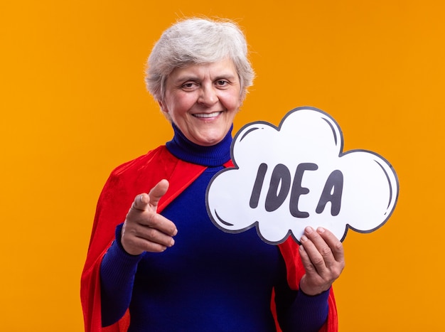 Senior woman superhero wearing red cape holding speech bubble sign with word idea pointing with index finger at camera happy and positive standing over orange background