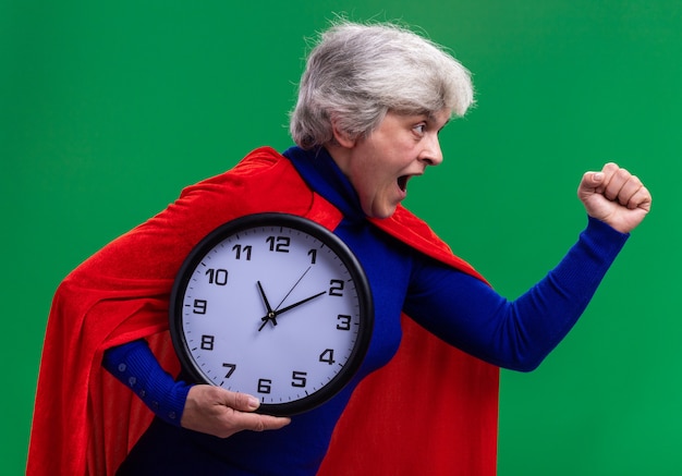 Senior woman superhero wearing red cape holding wall clock hurrying to help 