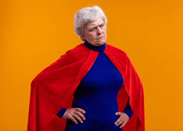 オレンジ色の背景の上に立っている悲しい表情で脇を見て赤いマントを身に着けている年配の女性のスーパーヒーロー