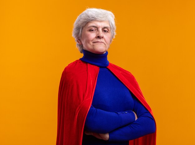 Senior woman superhero wearing red cape looking at camera with confident expression with arms crossed standing over orange background