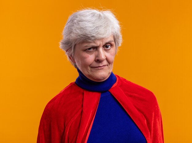 Senior woman superhero wearing red cape looking at camera with skeptic expression standing over orange background