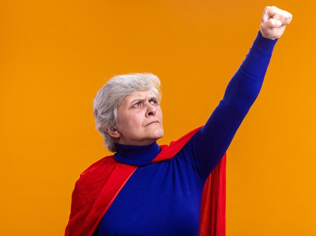 Старшая женщина-супергерой в красном плаще смотрит вверх, делая жест победителя с рукой, готовой помочь и бороться, стоя над оранжевым
