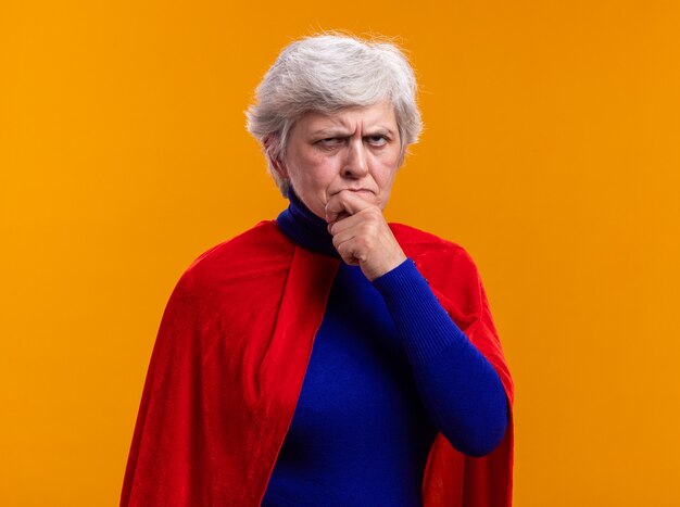 オレンジ色の背景の上に立っている眉をひそめている顔に困惑して見上げる赤いマントを身に着けている年配の女性のスーパーヒーロー