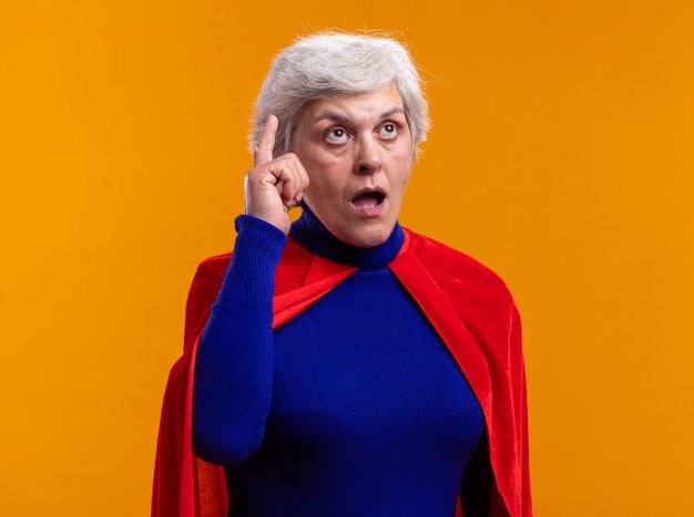 Senior woman superhero wearing red cape looking up showing index finger being surprised having new idea standing over orange