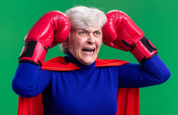 Senior woman superhero wearing red cape with boxing gloves frustrated and crazy mad touching her head standing over green background