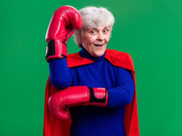 Senior woman superhero wearing red cape with boxing gloves looking at camera happy and surprised standing over green background